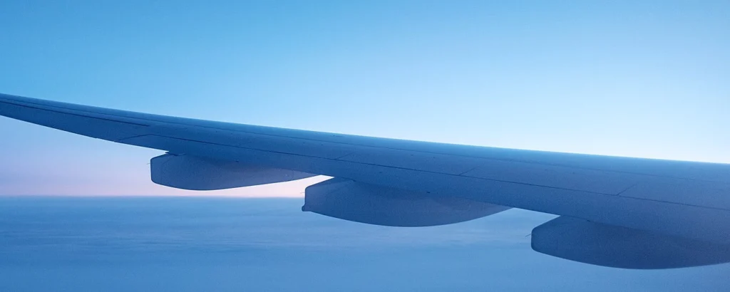 Aircraft wing above the clouds: the wing and engine nacelles against a pale blue sky and a smooth layer of cloud.