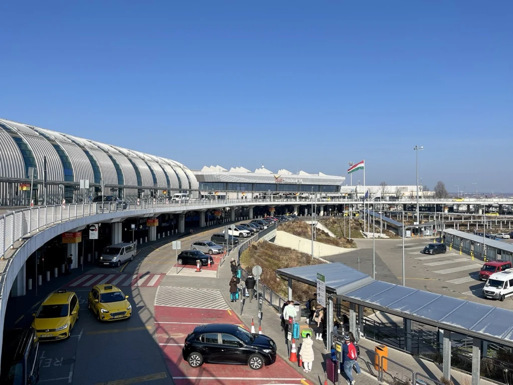 Terminal 2A of Budapest Liszt Ferenc Airport under a clear blue sky: a curved glass façade with taxis and passengers waiting in front, and a Hungarian flag in the background.